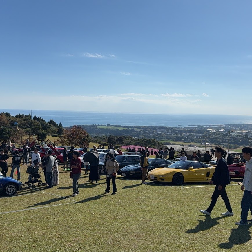 丘の上に旧車が並び、人々が集う旧車の祭典の会場。遠くに海が見える